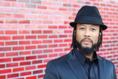 Siyah takım elbiseli, kırmızı tuğla duvarın önünde şapka ve rastalı genç adamın yakın plan portresi.