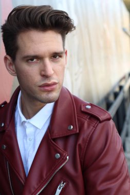 Bad boy concept. Portrait of brutal young man with short hair wearing red leather jacket, jeans and posing over urban background. Hand in pockets. Hipster style. Copy-space. Outdoor shot