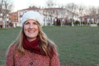 Kış giysileri içinde gülen sarışın kadının portresi gündüz vakti parkta duruyor.