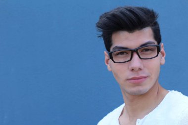 Mavi duvarın önünde duran beyaz gömlekli ve gözlüklü yakışıklı genç adamın yakın plan portresi.