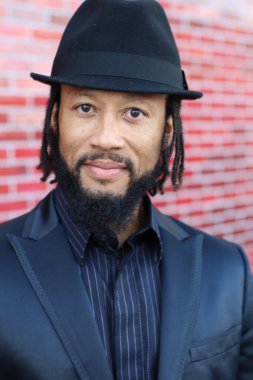 Siyah takım elbiseli, kırmızı tuğla duvarın önünde şapka ve rastalı genç adamın yakın plan portresi.