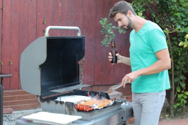 Genç sakallı adamın açık havada gündüz vakti bahçede BBQ 'da yemek pişirme portresi. 