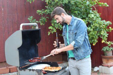 Genç sakallı adamın açık havada gündüz vakti bahçede BBQ 'da yemek pişirme portresi. 