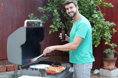 Genç sakallı adamın açık havada gündüz vakti bahçede BBQ 'da yemek pişirme portresi. 