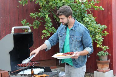 Genç sakallı adamın açık havada gündüz vakti bahçede BBQ 'da yemek pişirme portresi. 