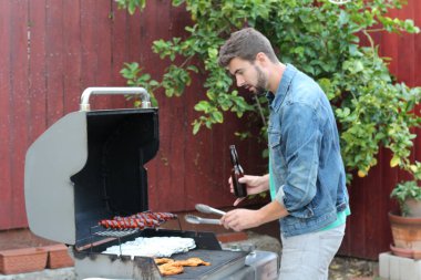 Genç sakallı adamın açık havada gündüz vakti bahçede BBQ 'da yemek pişirme portresi. 