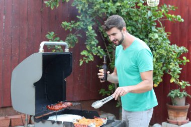 Genç sakallı adamın açık havada gündüz vakti bahçede BBQ 'da yemek pişirme portresi. 