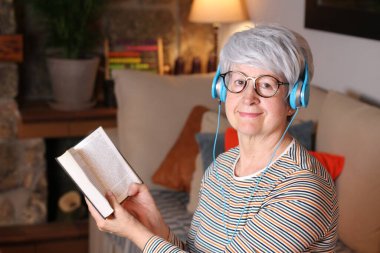 Karantina sırasında evde oturup kitap okuyan ve kulaklıkla müzik dinleyen olgun bir kadının yakın plan portresi.