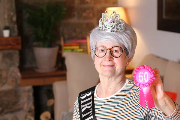 close-up portrait of mature woman with tiara, birthday stripe and 60 years birthday award sitting at home during quarantine