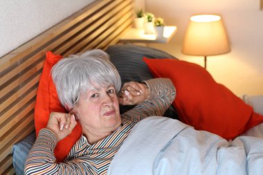 Olgun gri saçlı kadın evde yalnızlık çekerken parmaklarını kulaklara takıyor.