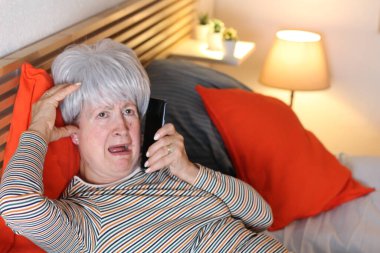 Olgun gri saçlı kadın evde yalnızlık çekerken yatakta dinlenirken telefonla konuşuyor.