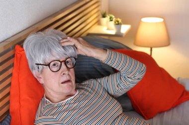 Olgun gri saçlı kadın evde yalnızlık çekerken yatakta dinlenirken baş ağrısı çekiyor.