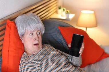 Olgun gri saçlı kadın evde yalnızlık çekerken yatakta dinlenirken boş ekranla akıllı telefon gösteriyor.