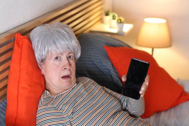 Olgun gri saçlı kadın evde yalnızlık çekerken yatakta dinlenirken boş ekranla akıllı telefon gösteriyor.