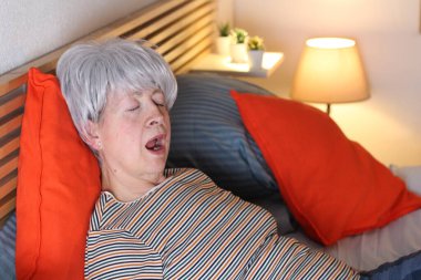 Olgun gri saçlı kadın evde yalnızlık çekerken yatakta esniyor.
