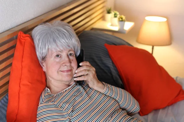 mature grey haired woman talking by phone while relaxing in bed during self isolation at home