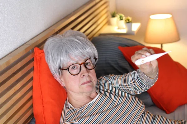 自宅で隔離中にベッドでリラックスしながら温度計を示す成熟した白髪の女性 — ストック写真