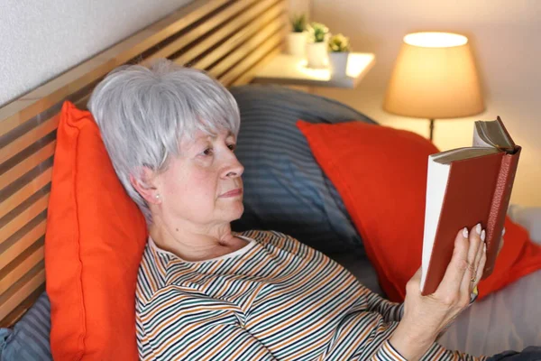 mature grey haired woman reading book while relaxing in bed during self isolation at home