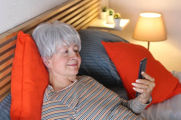 mature grey haired woman making video call with smartphone while relaxing in bed during self isolation at home