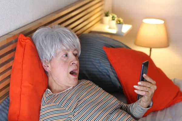 成熟的灰发女人在家里自我隔离时 一边在床上放松 一边用智能手机打视频电话 — 图库照片