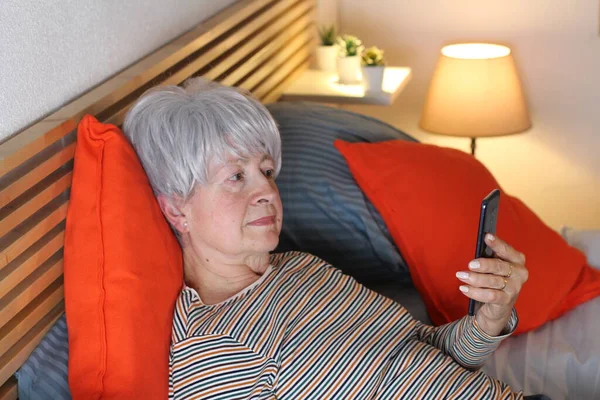 成熟的灰发女人在家里自我隔离时 一边在床上放松 一边用智能手机打视频电话 — 图库照片