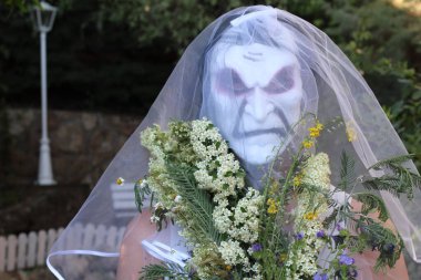 Gelinin korku maskeli portresi, gelin buketi ve yeşil bahçeli elbisesi.