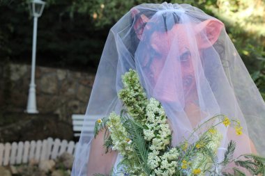 Gelinin korku maskeli portresi, gelin buketi ve yeşil bahçeli elbisesi.