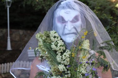 Gelinin korku maskeli portresi, gelin buketi ve yeşil bahçeli elbisesi.