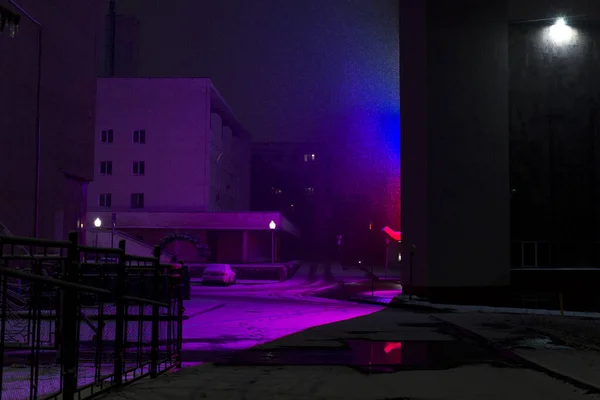 City buildings brightly lit in purple, red and blue, snowing in the light of lamps.