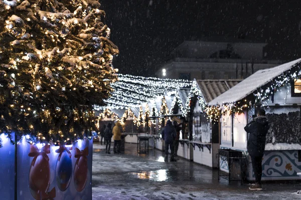 Şenlikli Noel ve Yeni Yıl panayırı geceleri yağan kar, yeni yıl ağacı ve çelenkli evler, yürüyen insanlar.