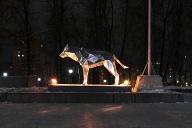 Şehir merkezindeki bir parkın ışıklarıyla aydınlatılan çokgen kurt metali figürü, şehir binalarının arka planında yer alıyor..