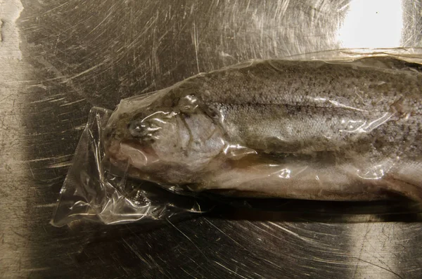 台所で木製のテーブルの上にプラスチック包装で密封され 冷凍ニジマス魚 調理の準備ができて — ストック写真