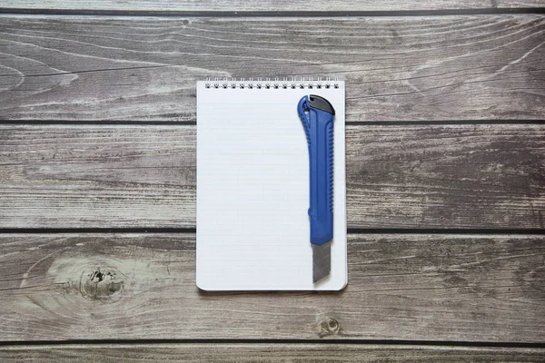 Bloc de notas con una hoja blanca en blanco en un papel de damero con cuchillo de papelería se encuentra en el fondo de las tablas de madera —  Fotos de Stock