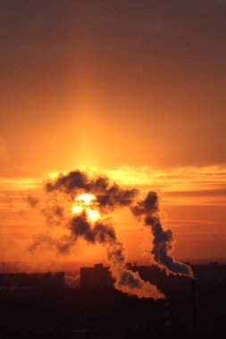 Red sun at sunset or dawn at dusk low above the horizon shrouded in smoke from the chimneys of the plant