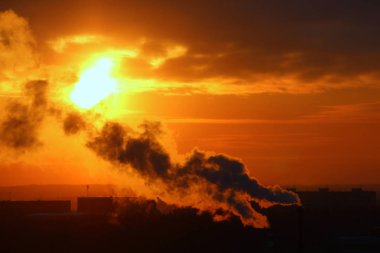 Red sun at sunset or dawn at dusk low above the horizon shrouded in smoke from the chimneys of the plant