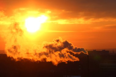 Red sun at sunset or dawn at dusk low above the horizon shrouded in smoke from the chimneys of the plant
