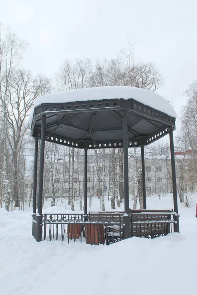 Metal gazebo aberto com bancos no parque, coberto com neve no dia nublado . — Fotografia de Stock