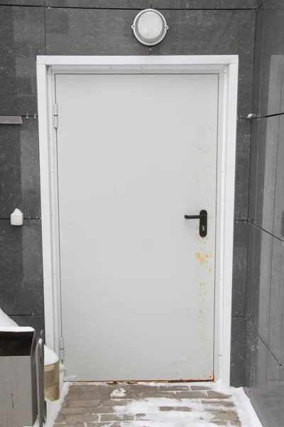 Белая входная дверь в серой облицованной панелями стене и покрытой снегом дороге . — стоковое фото