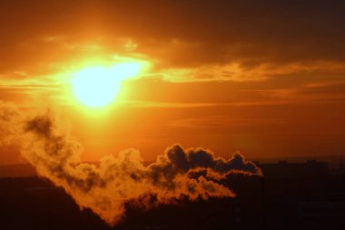 Red sun at sunset or dawn at dusk low above the horizon shrouded in smoke from the chimneys of the plant