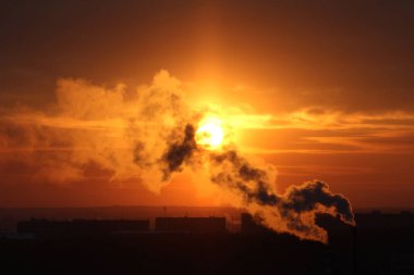Red sun at sunset or dawn at dusk low above the horizon shrouded in smoke from the chimneys of the plant