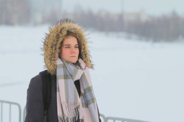 Güzel bir genç kız kış kıyafetleriyle dışarıda duruyor. Kış manzarasının arka planında bir ceket ve eşarp.