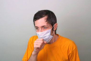 Orta yaşlı, sarı tişörtlü, gri arka planda tıbbi maske takmış yetişkin bir adam elleriyle yüzünü hapşırarak kapatıyor. Öksürük, ateş. Hastalıkları koruma Coronavirüs salgını Çin 'den geliyor..