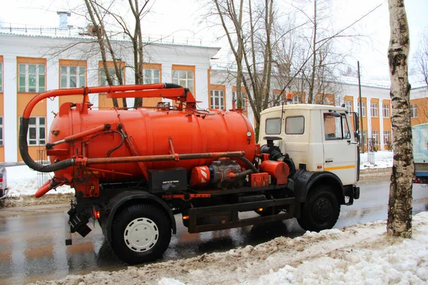 A truck with an orange tank with a white cab rides along a dirty city street in Russia in winter against the snow.