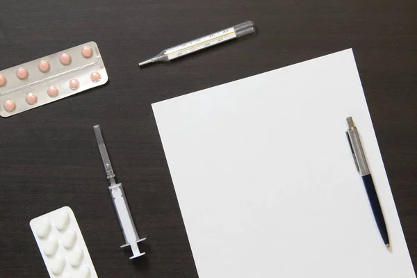 Template of white paper with pen, thermometer, syringe and pills in a blister on dark wenge color wooden background. The provision of medical and pharmacological services. With empty space for text.