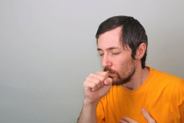 Middle-aged man with a mustache and a beard of a brunette in a yellow T-shirt on a gray background coughs, covering his mouth with his hand clenched in a fist and closing his eyes. clipart