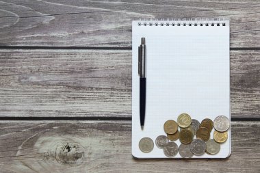 Dama kağıdında boş beyaz bir sayfa ve tahta tahtaların arka planında Rus sikkeleri ve tükenmez kalemleri olan bir not defteri var.