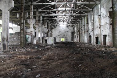 Terk edilmiş endüstriyel bir binanın içine bakın. Geniş bir alanı ve yüksek tavanı var. Sütunları ve tavandaki deliklerden gelen ışığı var. Zemin koyu kahverengi kumla kaplıdır..