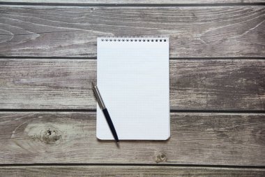 Notepad with a blank white sheet in a checker paper with ball pen lies on the background of wooden boards.