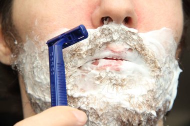 Bir adam mavi tek kullanımlık tıraş bıçağıyla sakalını kesiyor. Yanaklarında ve çenelerinde beyaz tıraş köpüğü.