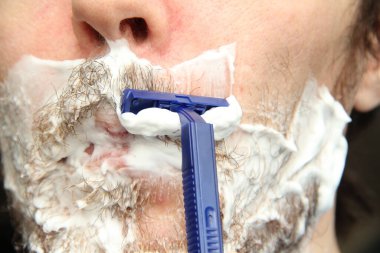 Bir adam mavi tek kullanımlık tıraş bıçağıyla sakalını kesiyor. Yanaklarında ve çenelerinde beyaz tıraş köpüğü.